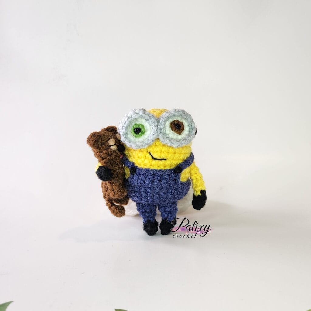 Bob minions - esp amigurumi, muñeco tejido con un osito, parado con un fondo blanco