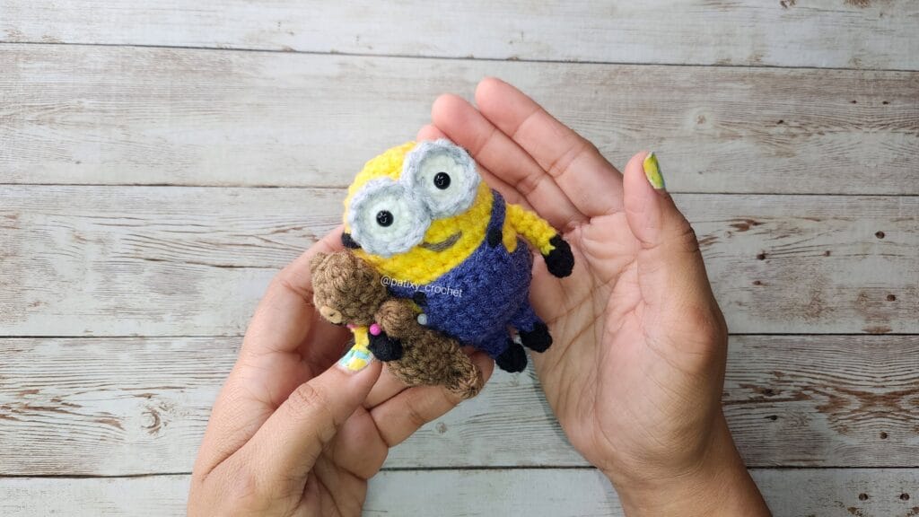 Bob minions - esp amigurumi, muñeco tejido con un osito sostenido con las manos sobre una mesa de madera blanca